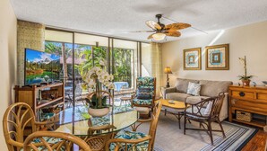 A spacious living area with a glass dining table, wicker chairs, a sofa, and a ceiling fan. - 🌴AC | Garden View | 1st floor | Kalanikai #110🌴 (Kailua Kona)
