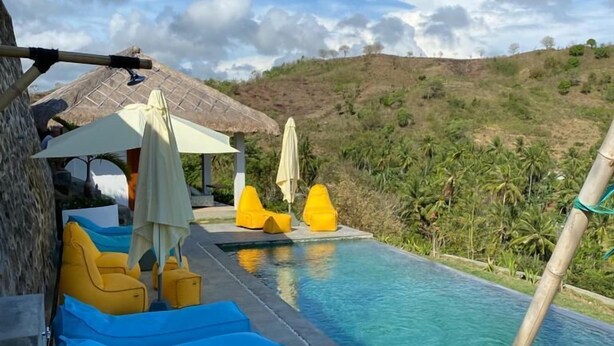 Uma área de piscina com cadeiras de descanso azuis e amarelas, uma estrutura com telhado de palha e vista para uma paisagem montanhosa.