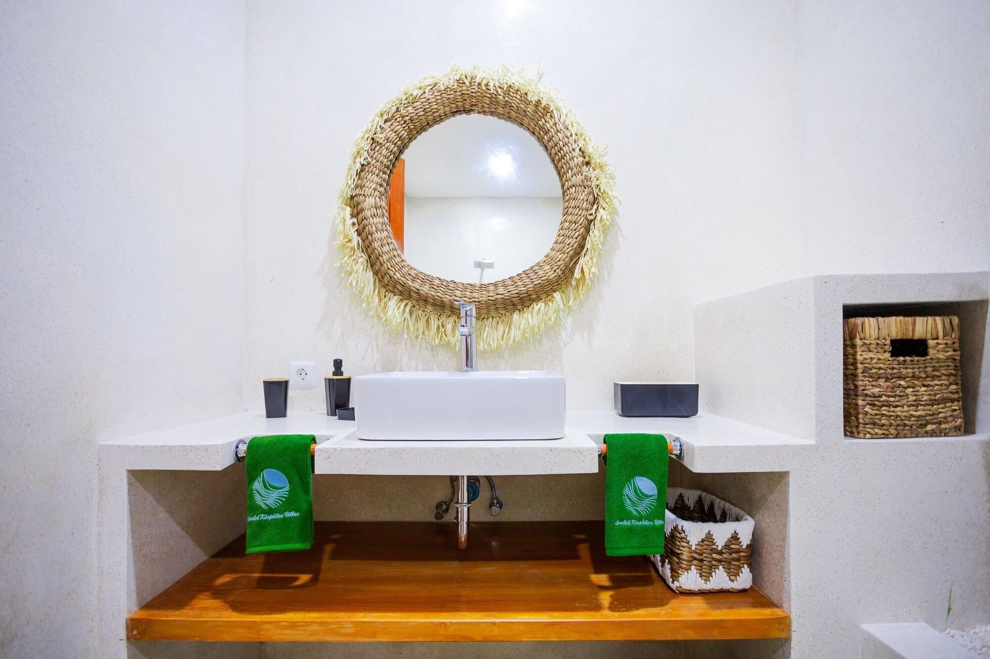 Een badkamer met een wit wastafel, twee groene handdoeken, een houten plank en een wanddecoratie van geweven materiaal.