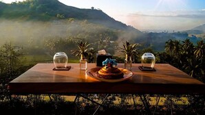 Uma mesa com bolo e copos, com vista para uma paisagem montanhosa e o sol.