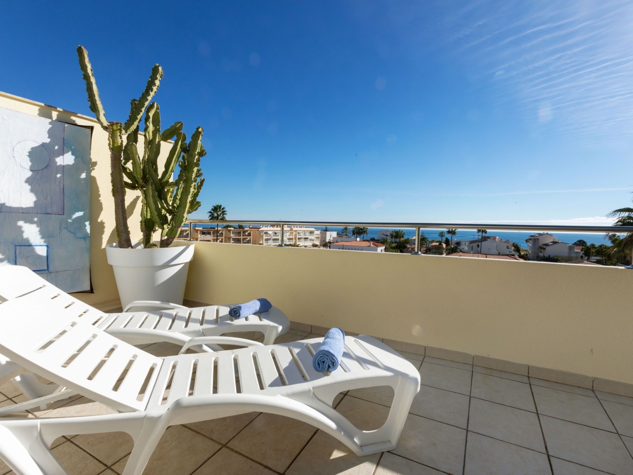 Ein Balkon mit weißen Plastikstühlen, einem Topf mit einem Kaktus und Blick auf das Meer und Gebäude.