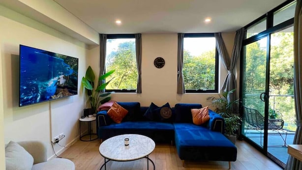 A modern living room with a sectional sofa, a flat-screen TV, a round coffee table, and large windows offering a view of greenery. - Majestic, Private Luxury Apartment - Harbourside (Darlinghurst)