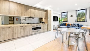 A modern kitchen with wooden cabinets, a central island, and a dining area with clear chairs. - Majestic, Private Luxury Apartment - Harbourside (Darlinghurst)