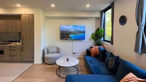 A modern living room with a sofa, a round coffee table, a TV mounted on the wall, and a kitchen area visible through an open door. - Majestic, Private Luxury Apartment - Harbourside (Darlinghurst)