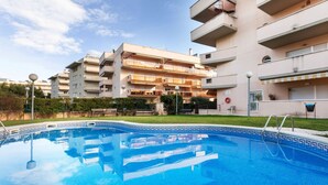 Une piscine dont le fond est recouvert de carreaux formant un motif géométrique, entourée d’une pelouse bien entretenue et d’un immeuble résidentiel à plusieurs étages doté de balcons.