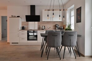 A modern kitchen with white cabinets, a black range hood, and a dining area with grey chairs.