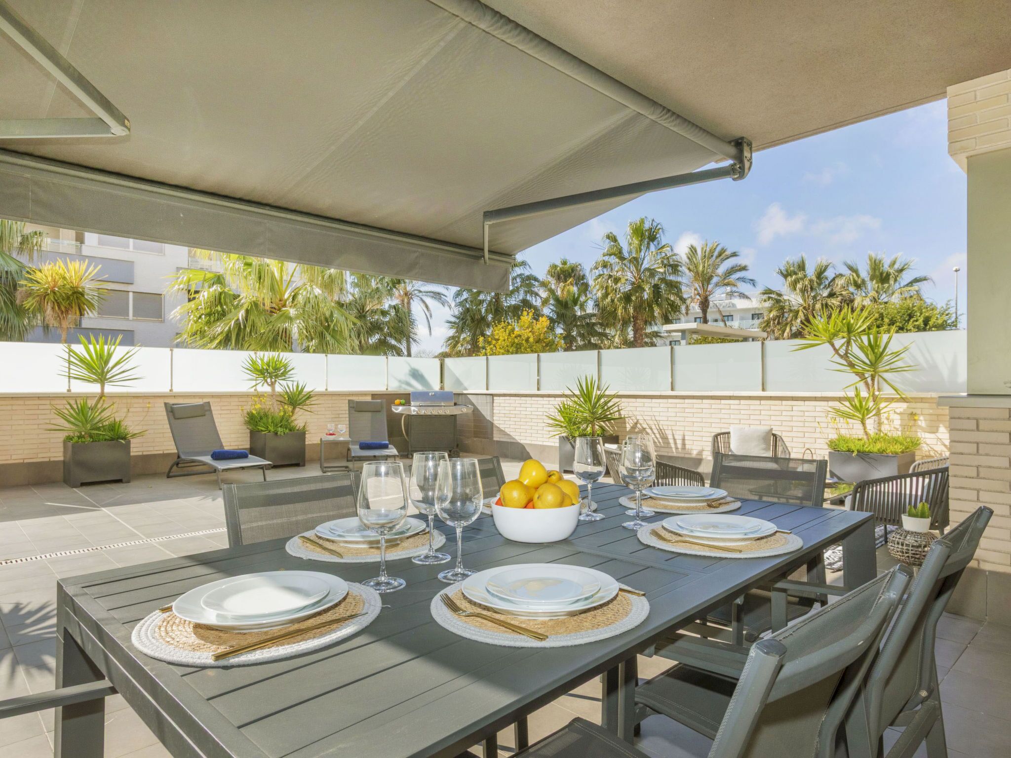 Un espace extérieur aménagé pour les repas, avec une table dressée pour six personnes, entourée de chaises, d’un barbecue et de plantes en pot.