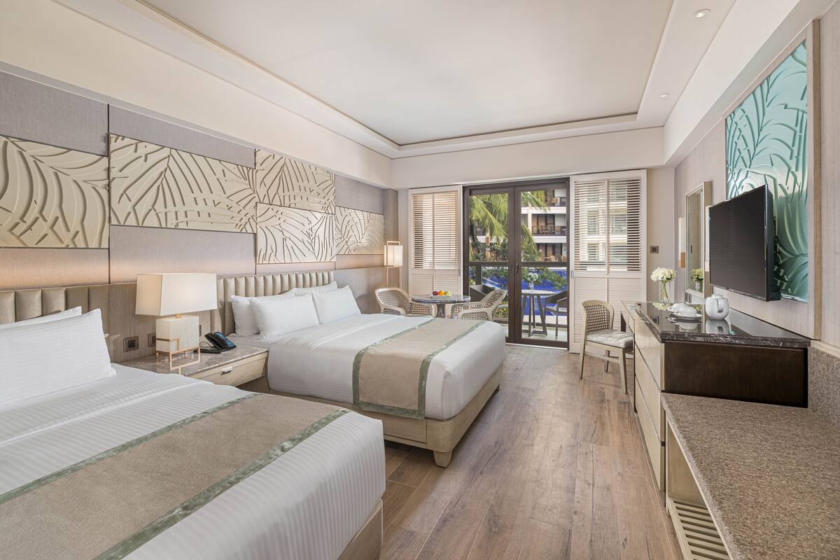 Habitación de hotel con dos camas, un escritorio, una silla, un televisor y un balcón con vistas a palmeras.