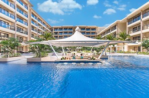 Zona de piscina de hotel con un área de estar techada y un edificio grande al fondo.