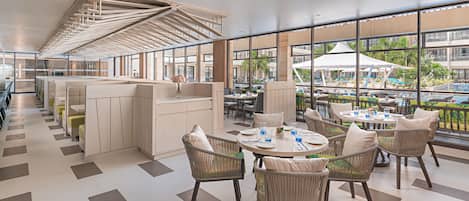 A modern hotel lobby with a high ceiling, open seating area, and large windows offering a view of the pool and outdoor area.
