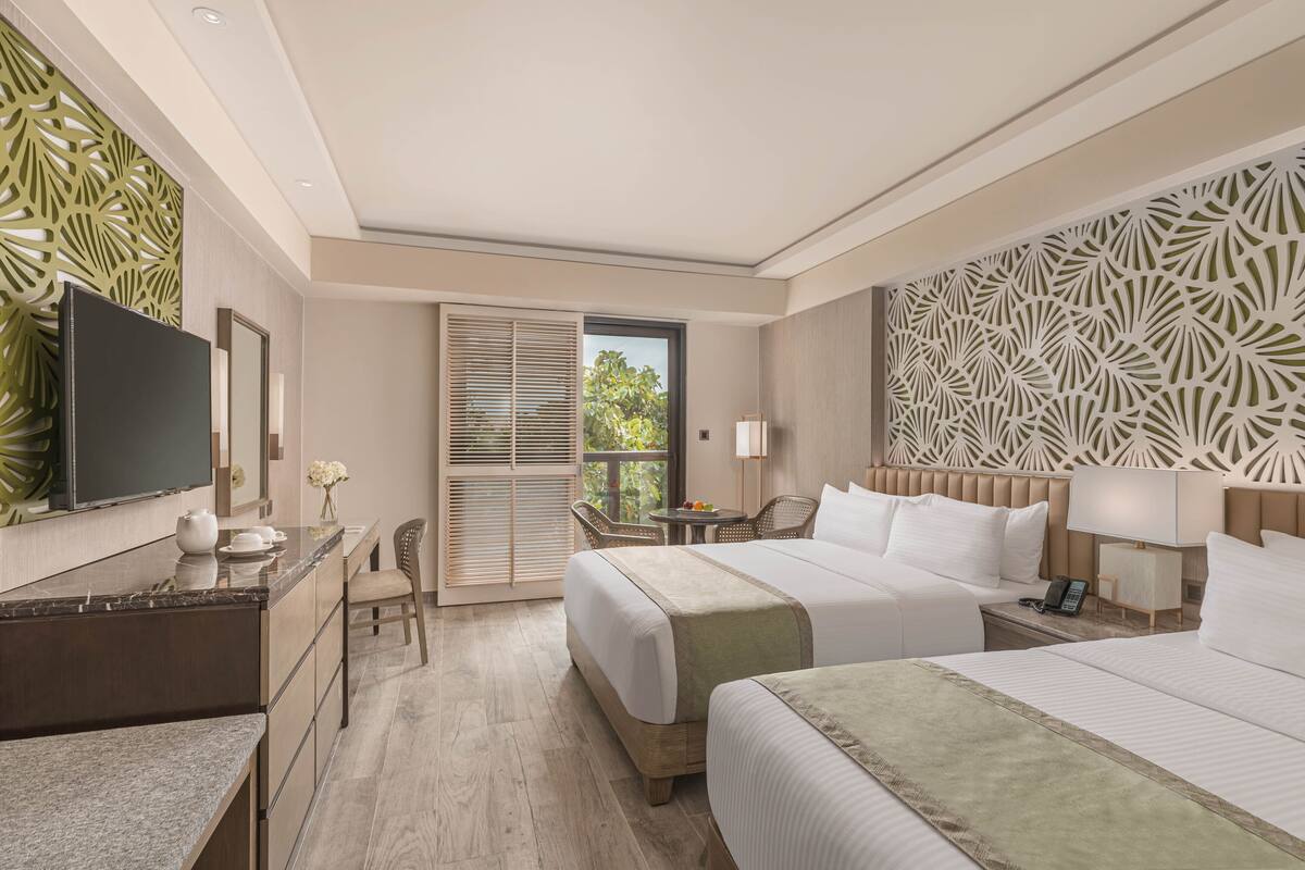 Habitación de hotel con una cama grande, un escritorio con televisor, una silla, una mesita con un florero y una ventana con persianas.