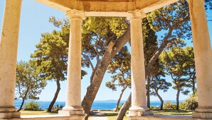 Un belvédère en pierre, doté de colonnes, entouré d’arbres et offrant une vue sur une étendue d’eau.