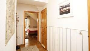 Ein schmaler Flur mit Holzboden, einem Bett mit roter Bettdecke, einer Holztür und einem gerahmten Bild an der Wand.