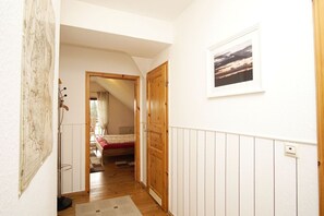 Ein schmaler Flur mit Holzboden, einem Bett mit roter Bettdecke, einer Holztür und einem gerahmten Bild an der Wand.
