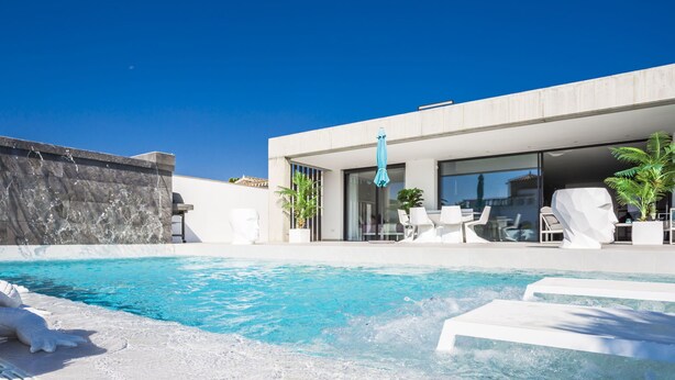 Un área de piscina moderna al aire libre, con una piscina de color azul cristalino, sillones de descanso blancos y un edificio contemporáneo con grandes ventanales.