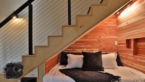 Une chambre avec un plafond en pente en bois, un lit avec des oreillers blancs et noirs, et un sac noir posé sur le sol.