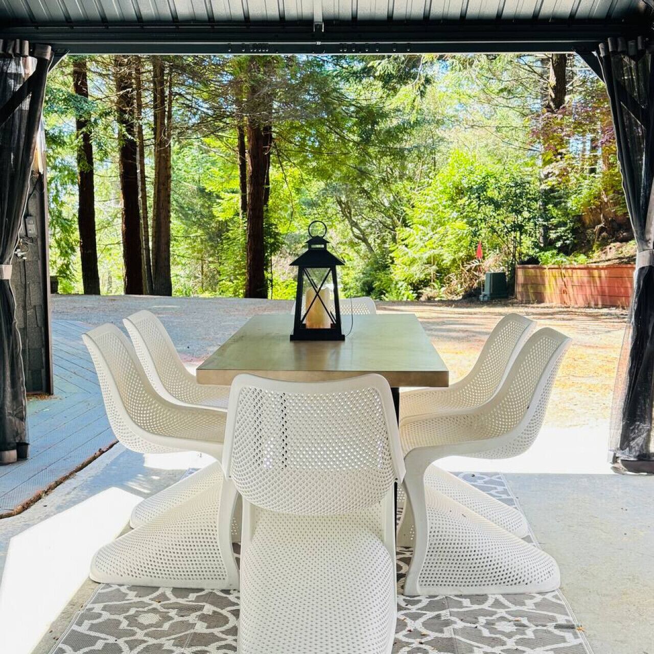 Un espace extérieur abrité, aménagé pour les repas, avec une table et six chaises blanches, le tout entouré d’une forêt.