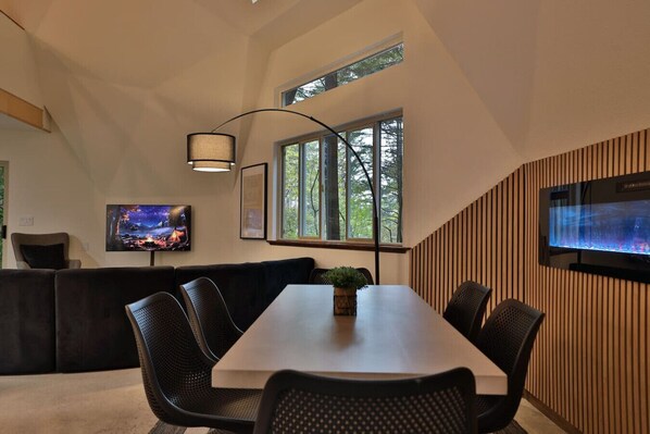 A modern dining area with a long table, black chairs, a sofa, a TV, and a large window with a view of trees.