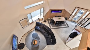 Un vaste espace de vie comprenant un canapé noir incurvé, une table basse ronde et une table à manger avec des chaises. De grandes fenêtres laissent entrer la lumière naturelle et offrent une vue sur l’extérieur.