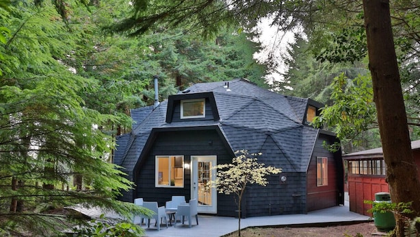 Une cabane à l’extérieur sombre, dotée d’un toit en pente et d’une terrasse avec des chaises.