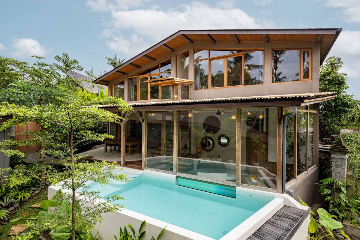 A modern two-story house with a glass-enclosed swimming pool, surrounded by lush greenery.