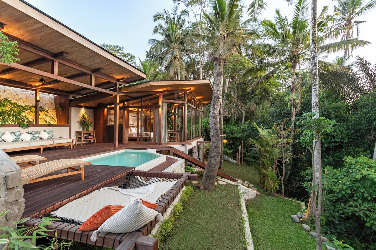 A poolside area with wooden deck seating, a stone wall, and lush tropical vegetation.