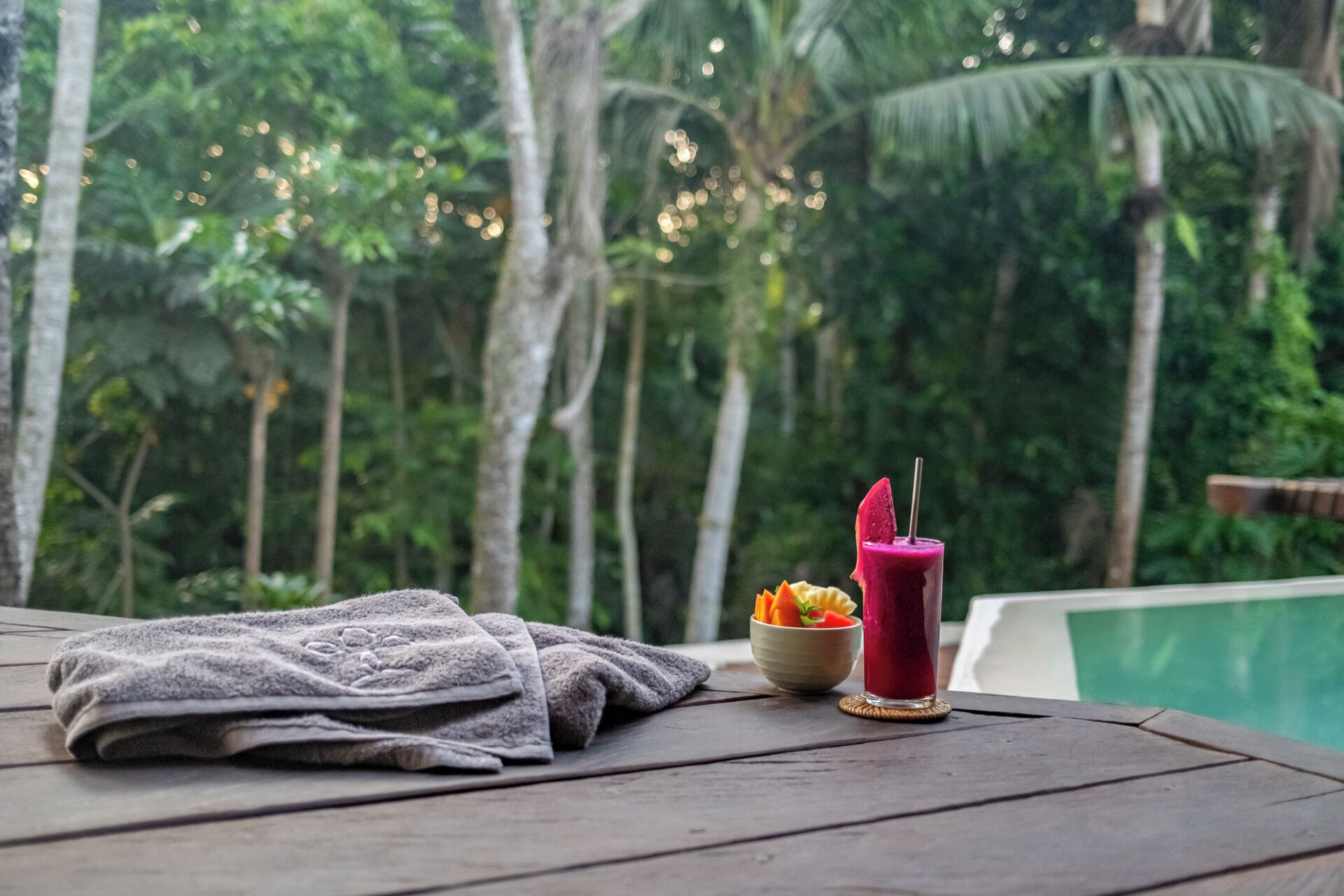 A wooden table with a folded towel, a bowl of fruit, and a glass of red drink.