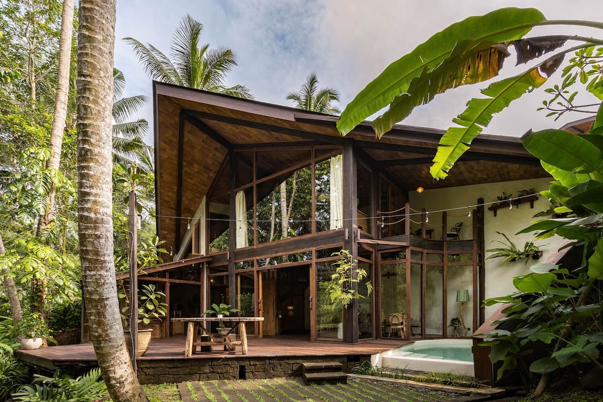 A modern house with a wooden deck, a glass-enclosed patio, and a small swimming pool surrounded by lush greenery.