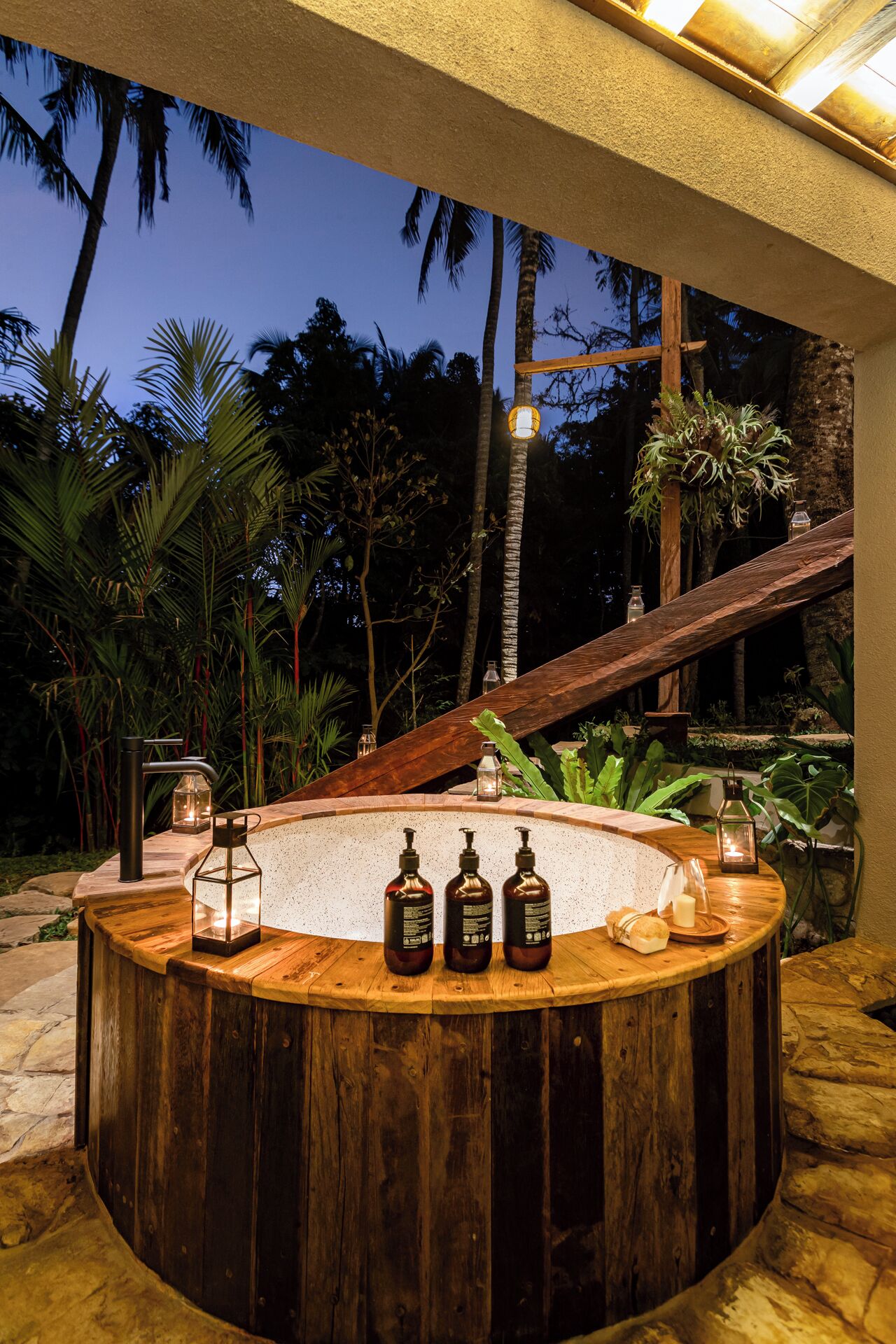 A wooden bar with a stone countertop, surrounded by tropical plants and a staircase leading to an upper level.