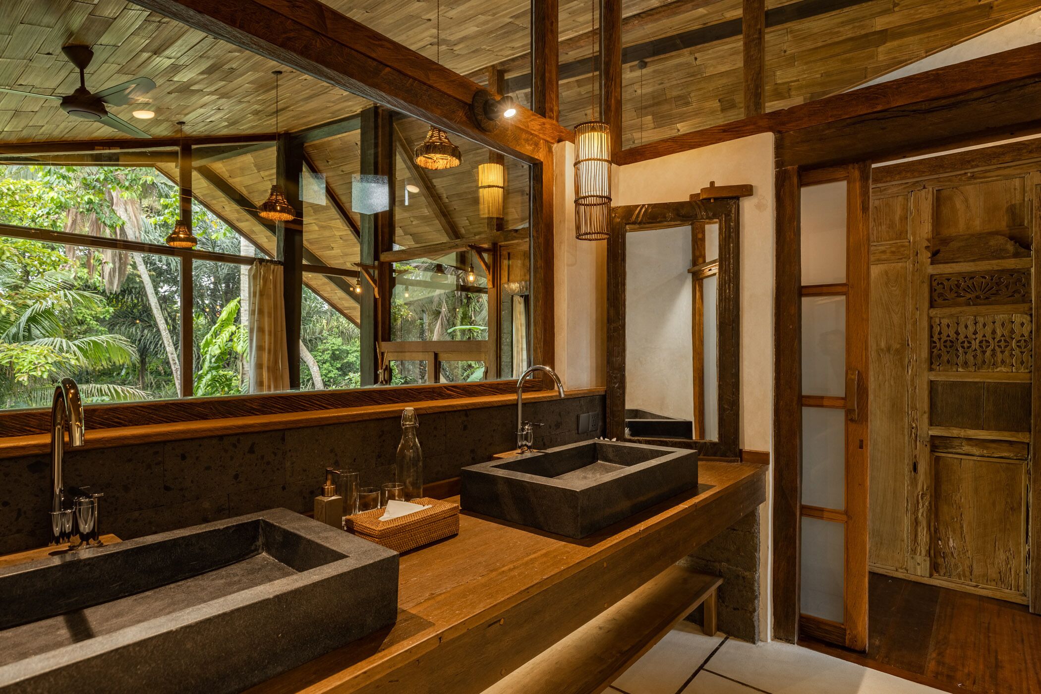A bathroom with a large mirror, two sinks, wooden beams, and a view of greenery outside.