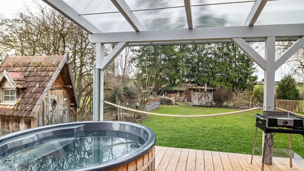 Una terraza de madera con un jacuzzi y una parrilla, rodeada por un patio cercado y árboles.