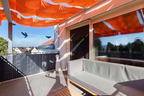An outdoor balcony with a seating area, a sliding glass door, and a view of the surrounding neighborhood.