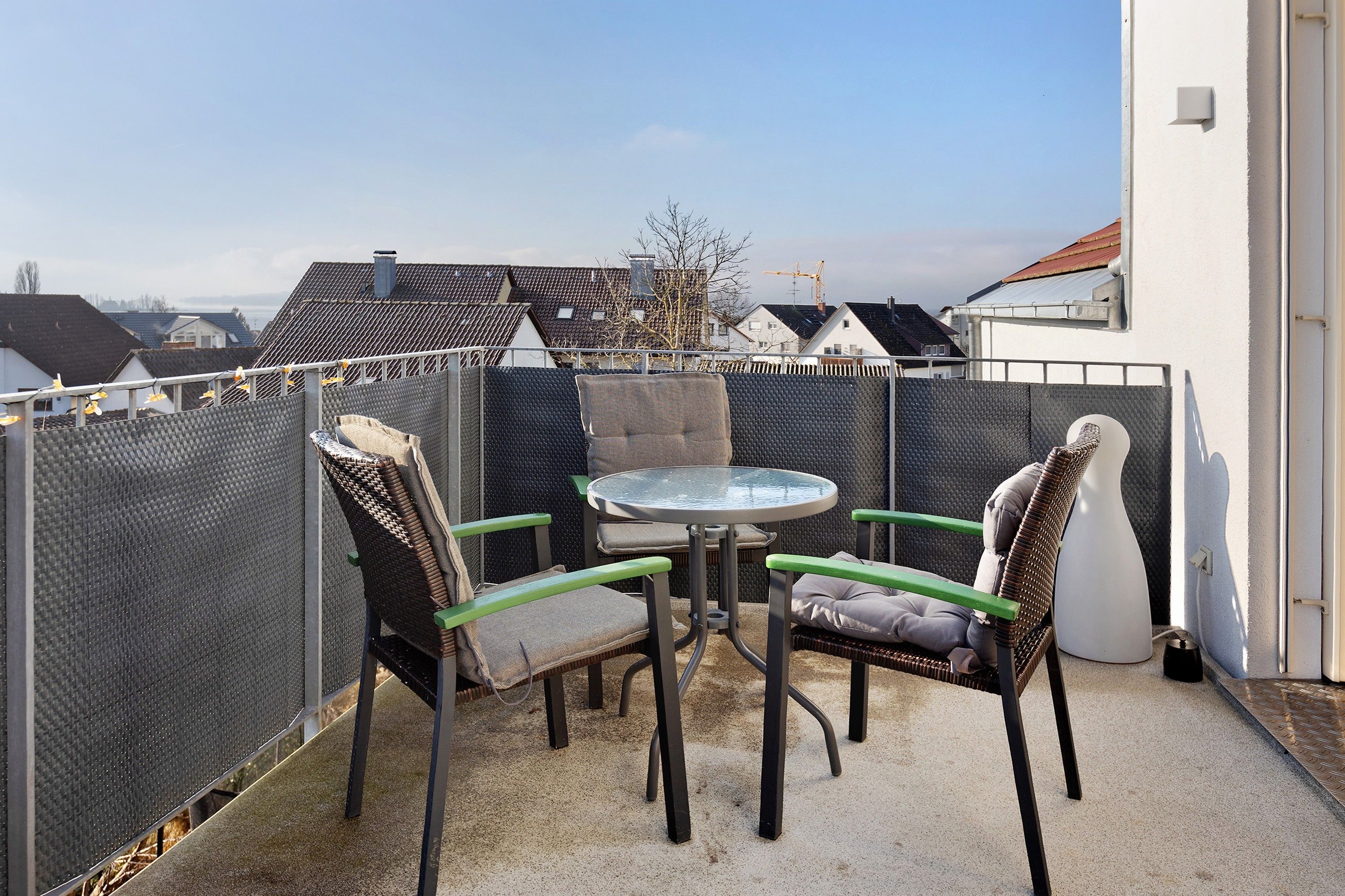 Ein Balkon mit rundem Tisch und zwei Stühlen, eine weiße Wand und Blick auf Gebäude im Hintergrund.