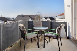 Ein Balkon mit rundem Tisch und zwei Stühlen, eine weiße Wand und Blick auf Gebäude im Hintergrund.