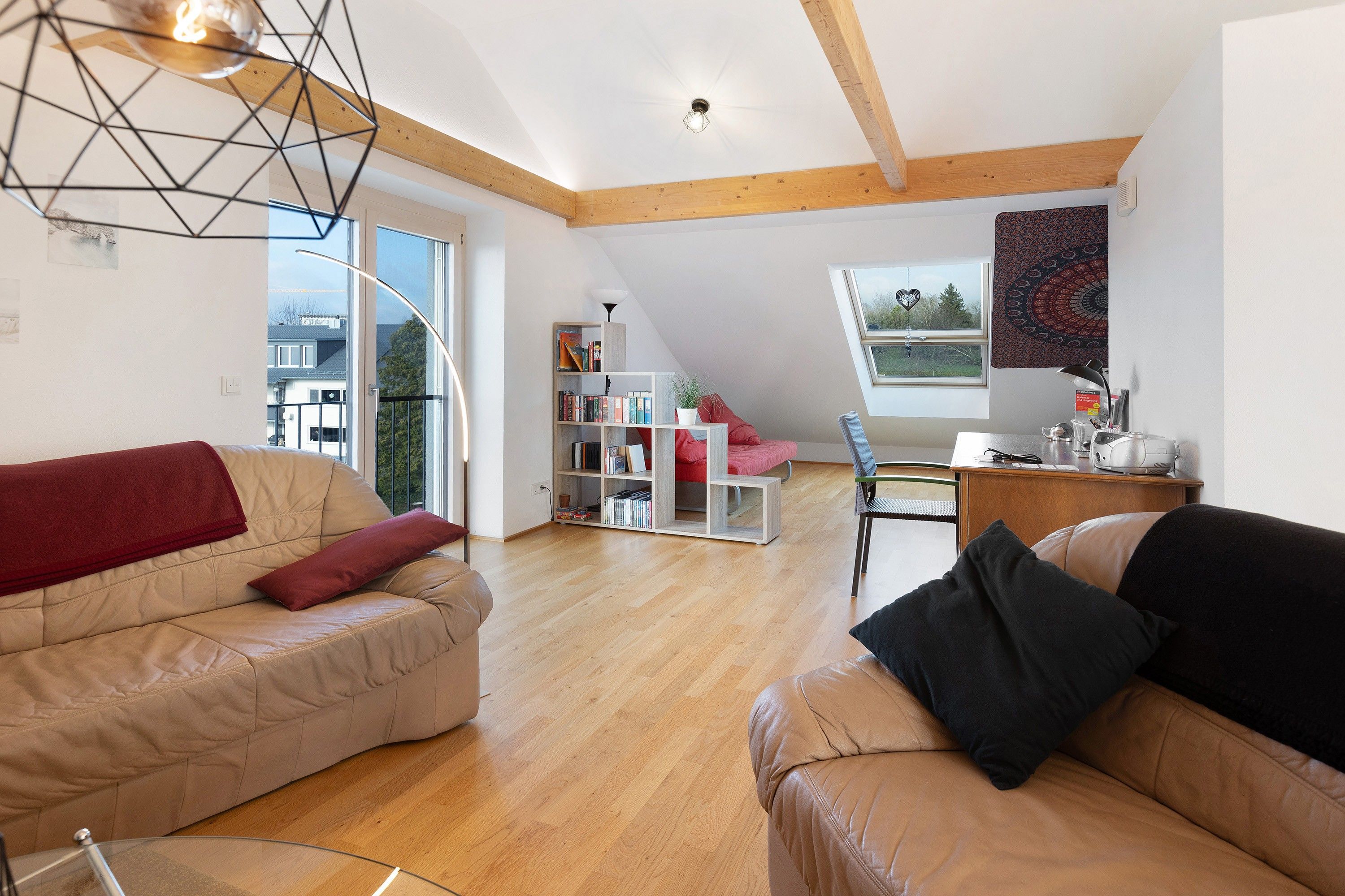Geräumiges Loft mit hohen Decken, Holzbalken, einem großen Fenster, einem roten Sofa, einem schwarzen Sofa und einem Bücherregal.