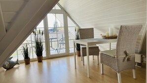 Un comedor con una mesa blanca, sillas de mimbre, ventanas amplias y plantas en macetas.