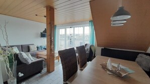 Een moderne woonkamer met een houten plafond, een eethoek met tafel en stoelen, een zithoek en een groot raam met gordijnen.