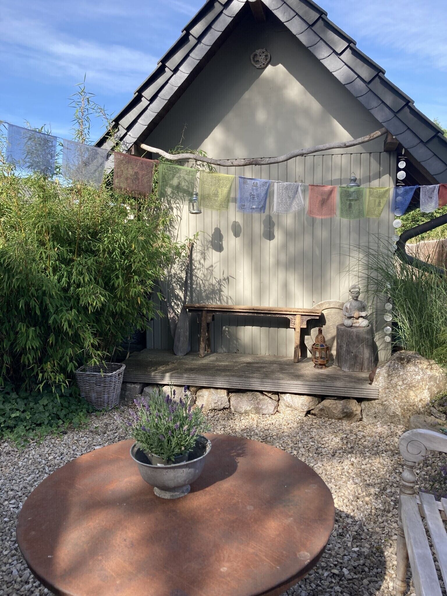 Eine rustikale Holzhütte mit einer Bank und einer Laterne, umgeben von Grünpflanzen und einem kleinen Tisch mit einem Blumentopf.