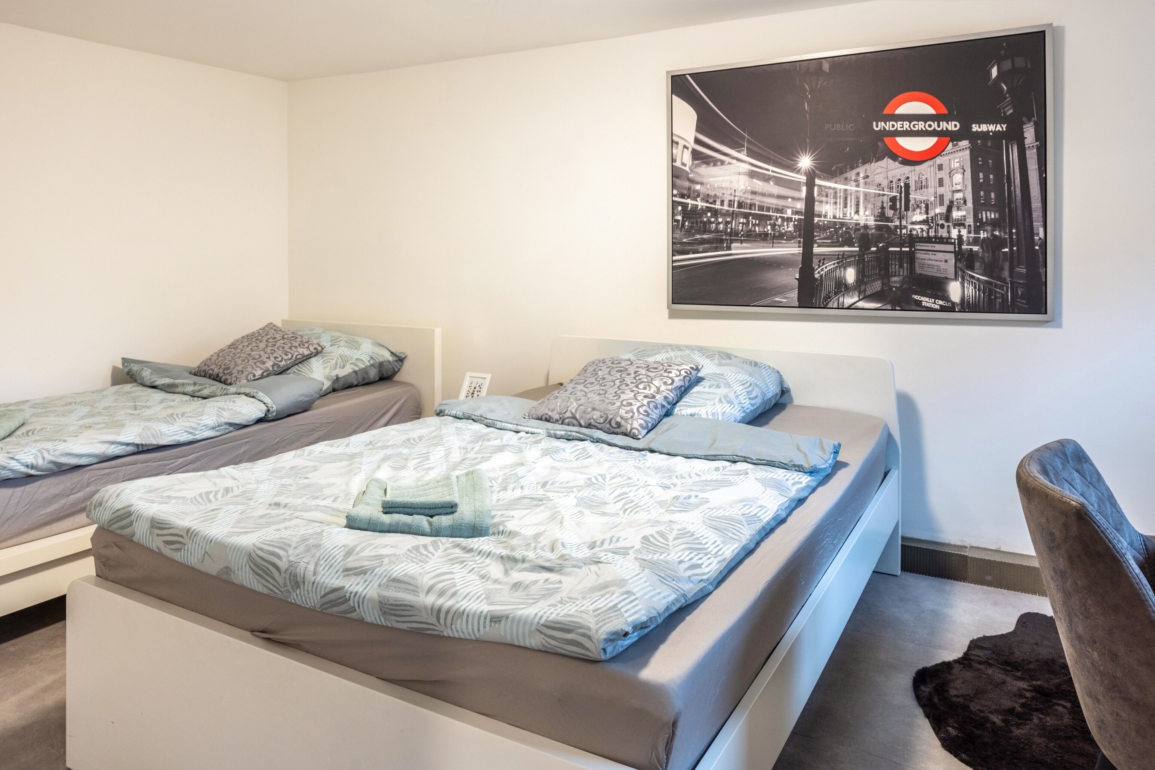 A hotel room with two beds, a brown chair, a poster of the London Underground, and a small rug.