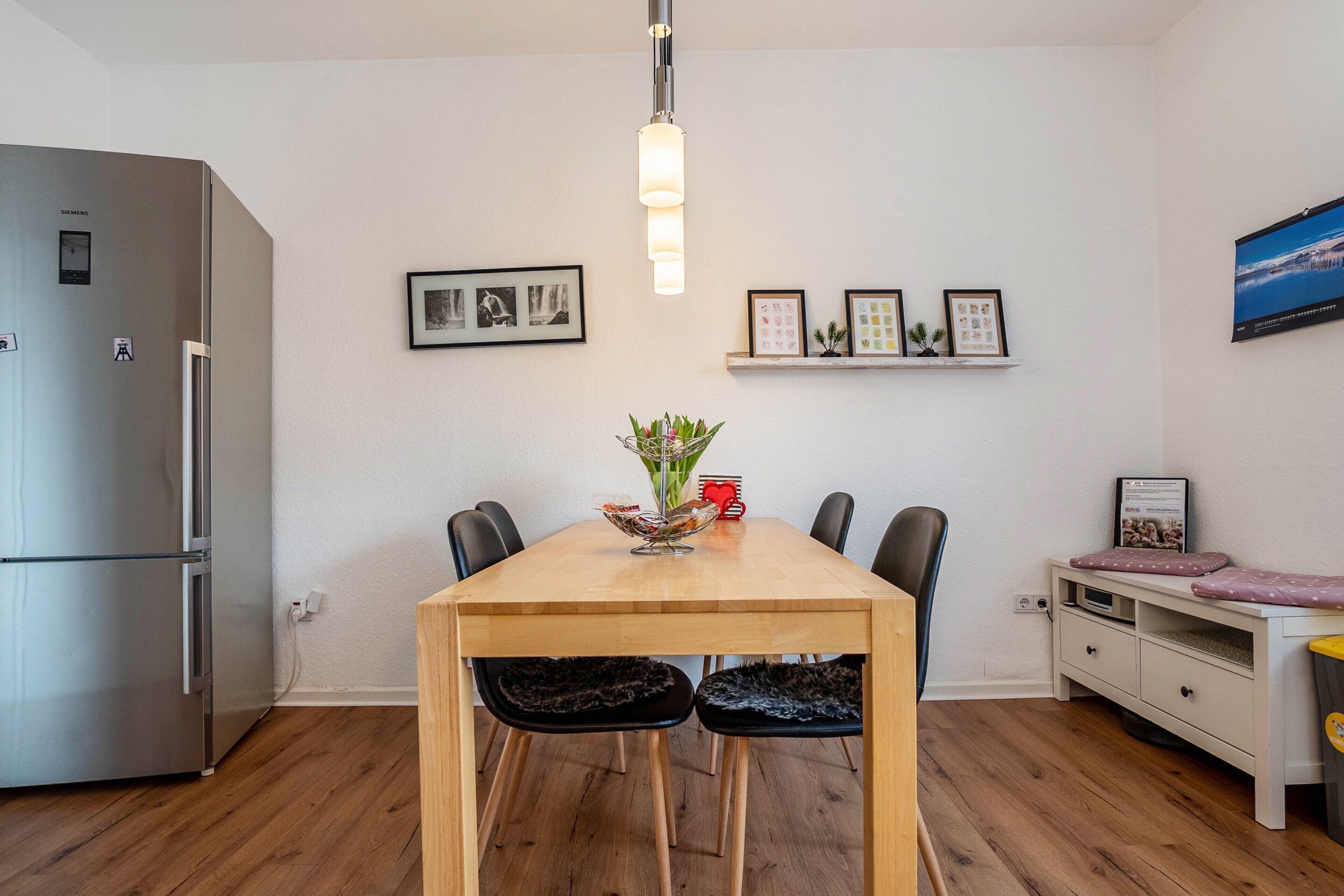 Eine moderne Küche mit Holztisch, Stühlen, einem Kühlschrank und an Wänden befestigten Regalen mit gerahmten Bildern.