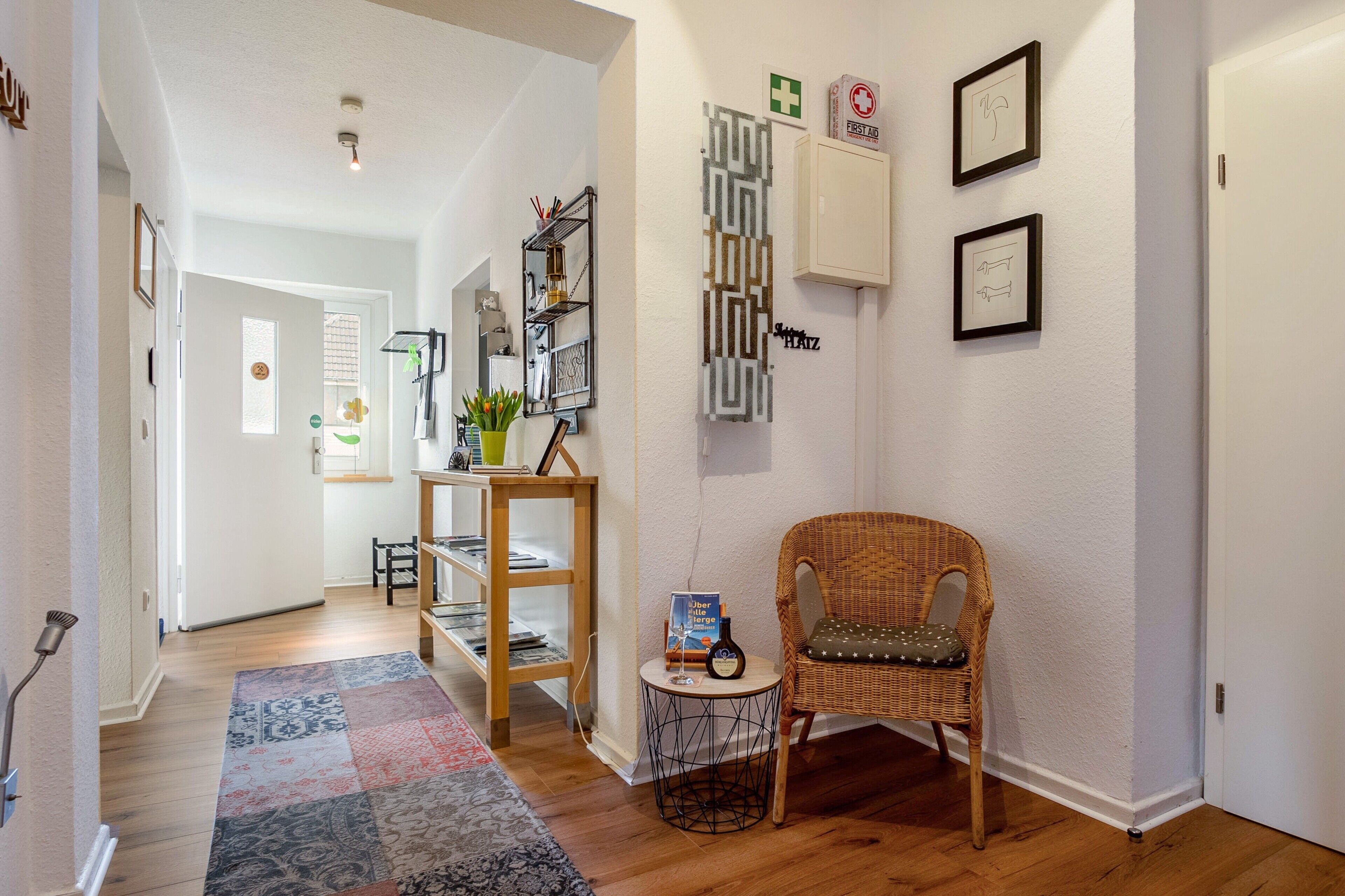 A hallway with a wicker chair, a small table with a book, a shelf with books, and a wall-mounted cabinet.