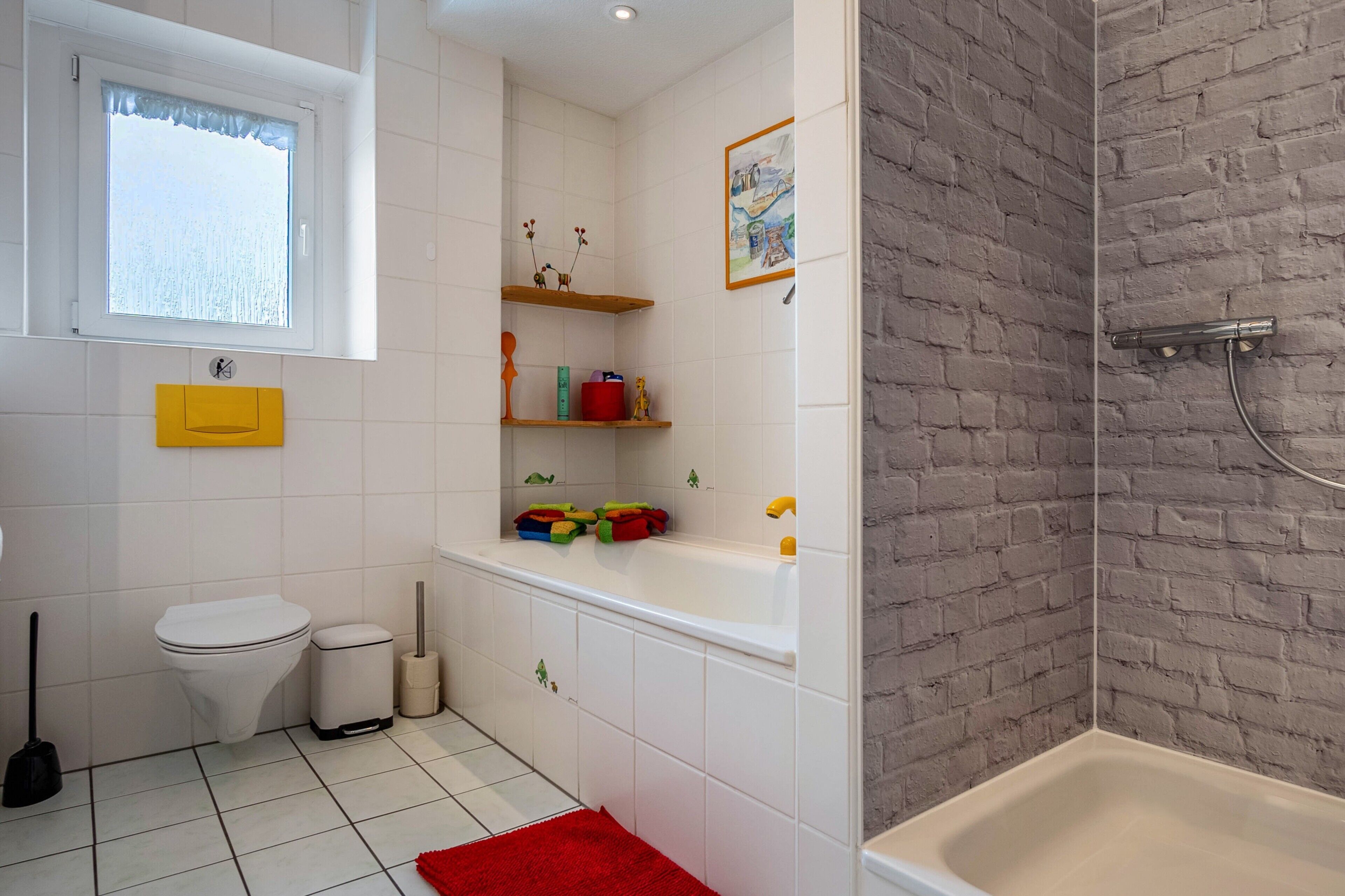 Een badkamer met een wit toilet, een wastafel, een badkuip en een douche. Er zijn planken met toiletartikelen en een raam boven het toilet.