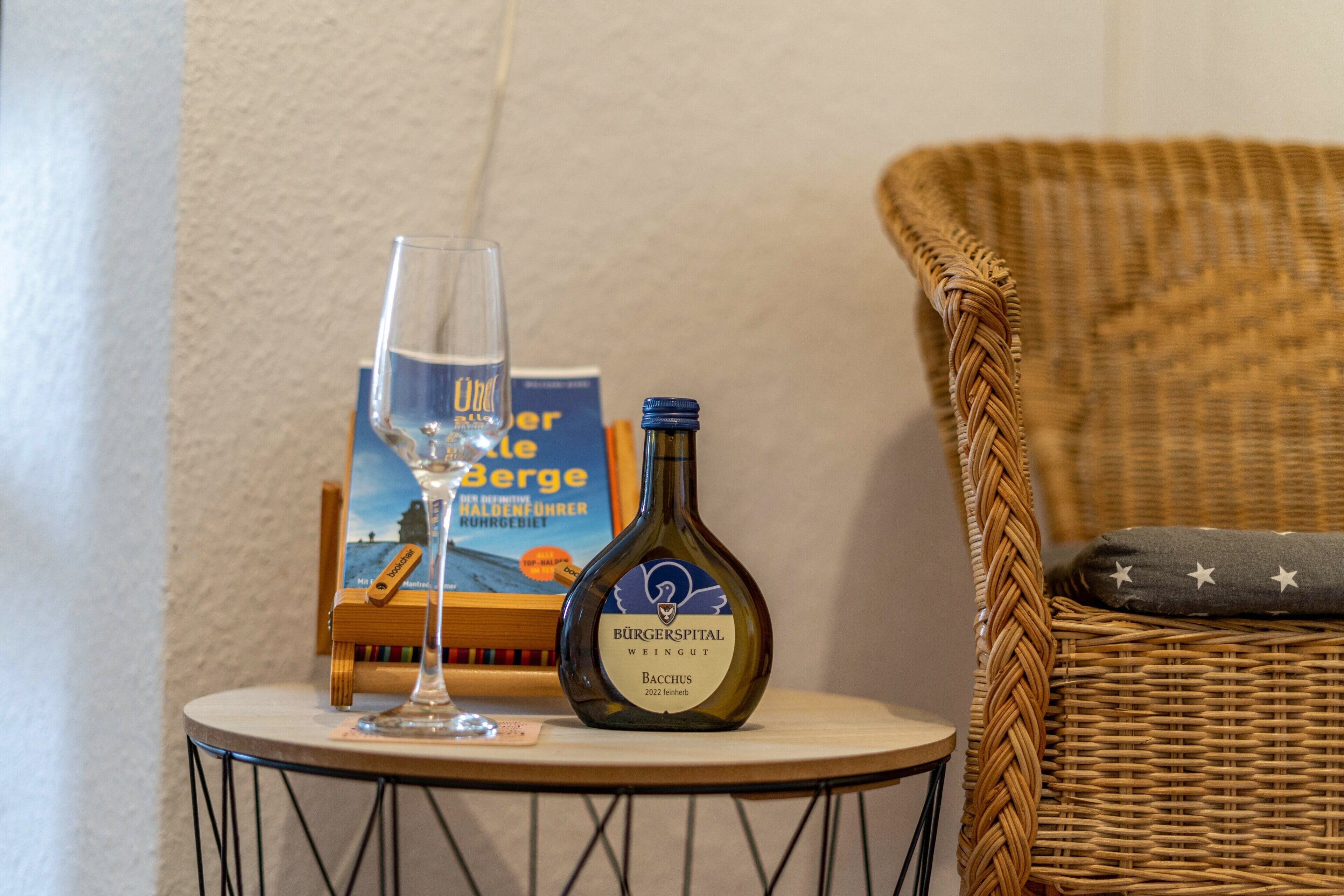 A bottle of Bäcchus wine, a glass of wine, and a book titled 'Burgerspital Weingut' on a small round table.
