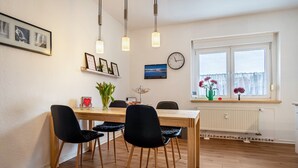 A dining area with a wooden table and four chairs, a clock, framed pictures, and a window with curtains. - Apartment 'Glückauf' with Wi-Fi (Castrop-Rauxel)