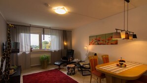 A living room with a TV, a dining table with fruit, a red rug, and a painting on the wall.