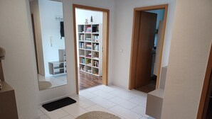 Een modern interieur met een vloer van witte tegels, een houten deur die leidt naar een kamer met een boekenkast en een spiegel die een deel van de ruimte weerspiegelt.