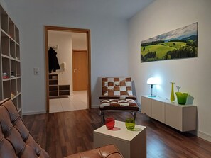 Un salon avec un canapé en cuir marron, un fauteuil à carreaux, une petite table blanche ornée de deux vases en verre et un tableau représentant un paysage accroché au mur.