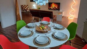 Een eetruimte met een ronde tafel voor zes personen, omringd door kleurrijke stoelen, een moderne hanglamp en een televisie waarop een haard te zien is.