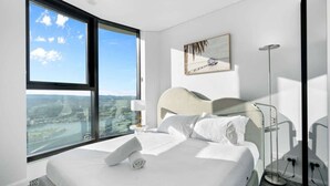Habitación de hotel con un ventanal amplio que ofrece vistas a la ciudad, una cama con sábanas blancas, un cabecero, una mesita de noche con lámpara y una lámpara de pie.