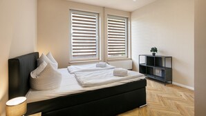 Un dormitorio moderno con cama negra, ropa de cama blanca, una lámpara de noche, suelo de madera y una mesita de noche negra con una planta.